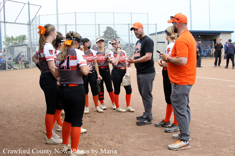 Softball player huddle: girls in orange socks and uniforms listen to two coaches on a dirt field, with a chain-link fence and dugout in the background.