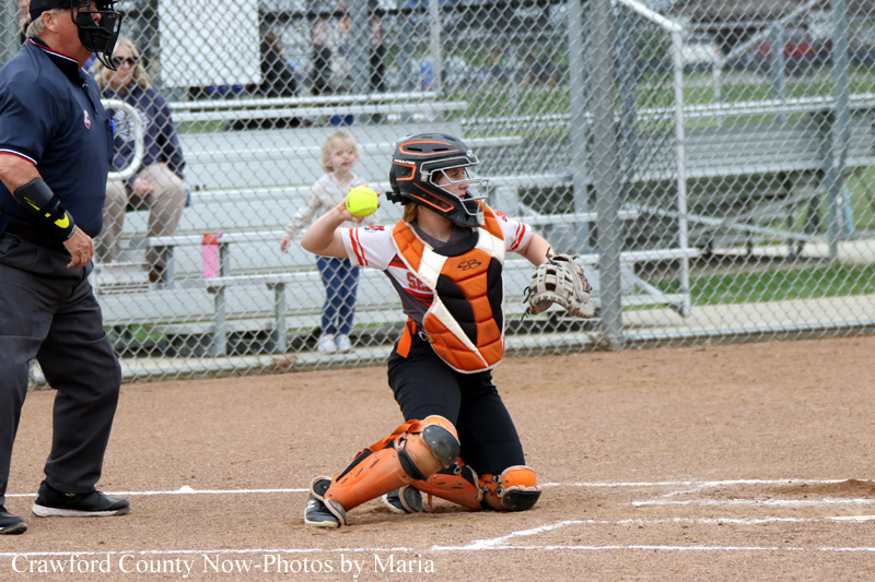 Youth softball catcher in orange and black protective gear kneels and throws a ball during a game, umpire and bleachers in background.