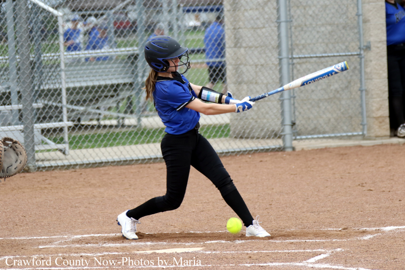 Young girl in a blue softball jersey swinging a bat at a yellow ball on a dirt field, wearing a helmet and gloves with a chain-link fence behind her.