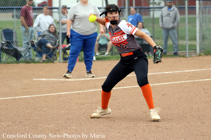 Female softball pitcher in mid-throw on the infield, glove up, bright orange socks, gray-orange uniform, batter’s box visible behind her.