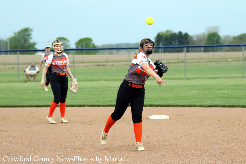 Softball pitcher in gray-red uniform winds up to throw on the infield; two teammates watch nearby in orange socks and masks.]”,