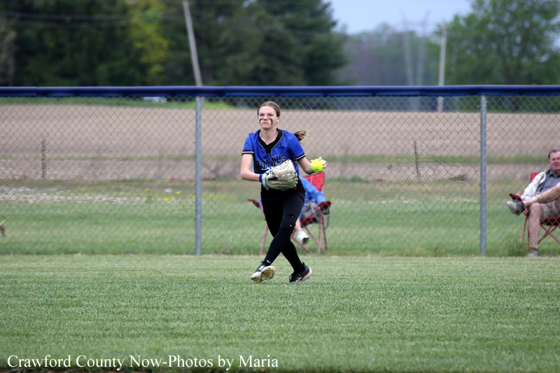 Female softball pitcher in a blue uniform mid-throw on a grassy field, with a chain-link fence and spectators in chairs behind her.