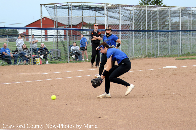 Female softball fielder in blue crouches to field a ground ball on the dirt infield while spectators watch behind the chain-link fence.