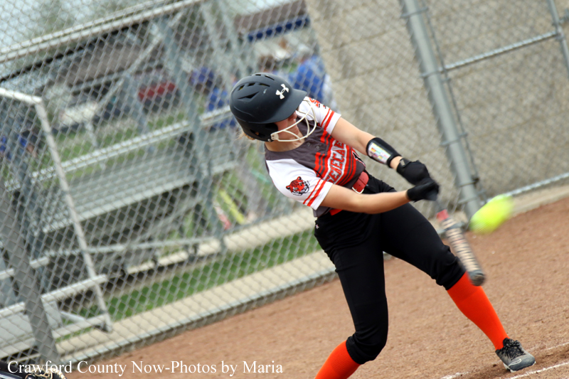 Female softball player in black pants and red socks swings a bat during a game, wearing a navy helmet and white-red jersey behind a chain-link fence.