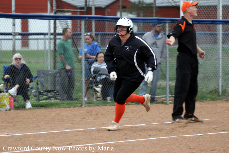 Female softball player in black uniform and white helmet sprints along the dirt basepath with orange socks, near an umpire and fence-lined field.