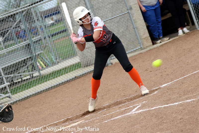 Female softball batter in a white helmet, white-and-black uniform, and orange socks swings at a pitch on a dirt field as the ball approaches.