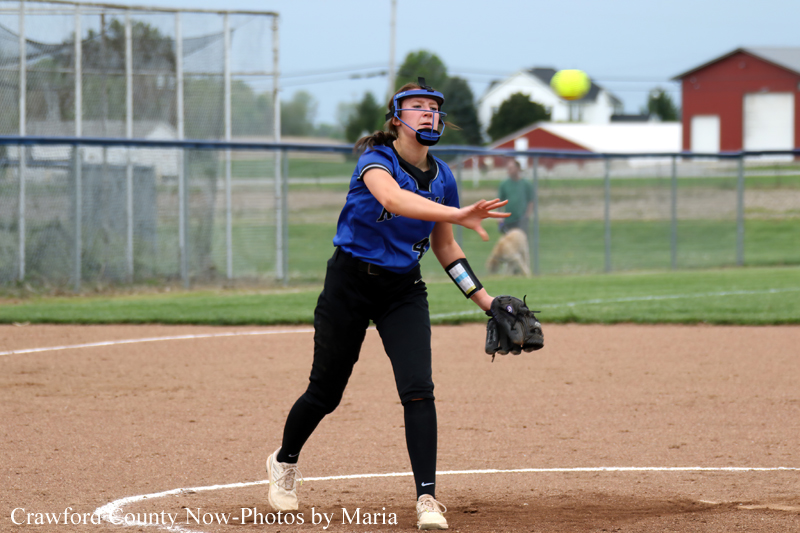 Female softball pitcher in a blue uniform throws a pitch on the dirt infield, wearing a protective face mask and glove on her left hand.