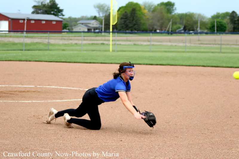 Female softball player in a blue shirt and mask dives on the infield with glove reaching for a ball