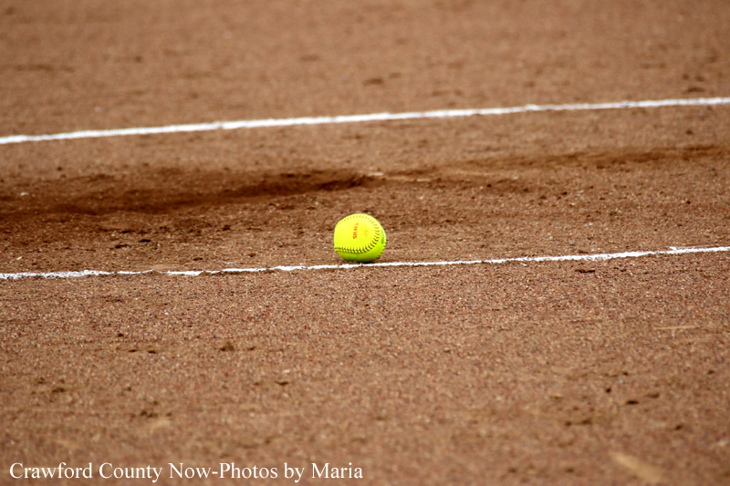 Bright yellow softball resting on a brown dirt infield with white chalk lines nearby.