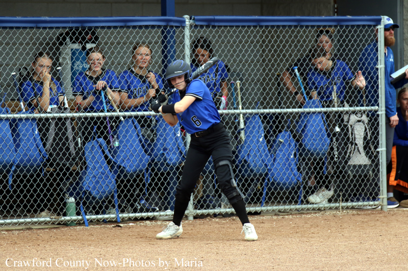 Baseball batter in a blue uniform stands in the batter's box, helmet on, gripping the bat as teammates watch from behind a chain-link fence.