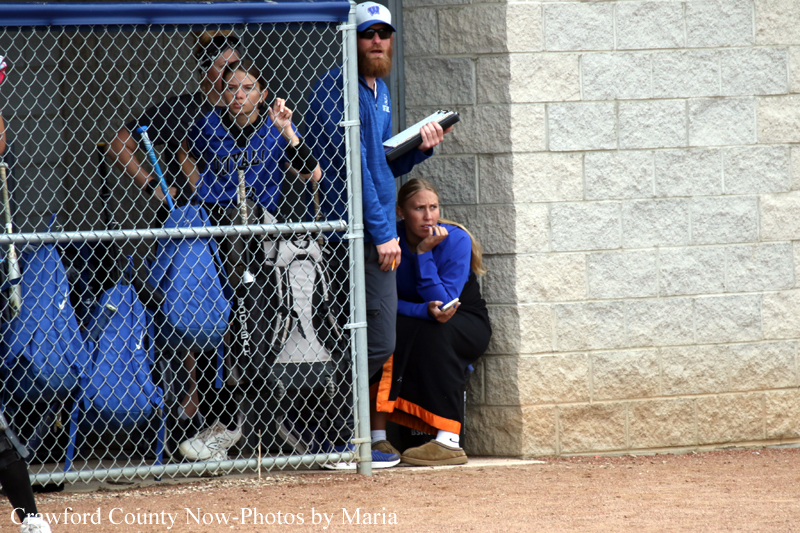 Baseball players in blue uniforms inside a chain-link dugout, watching the game.