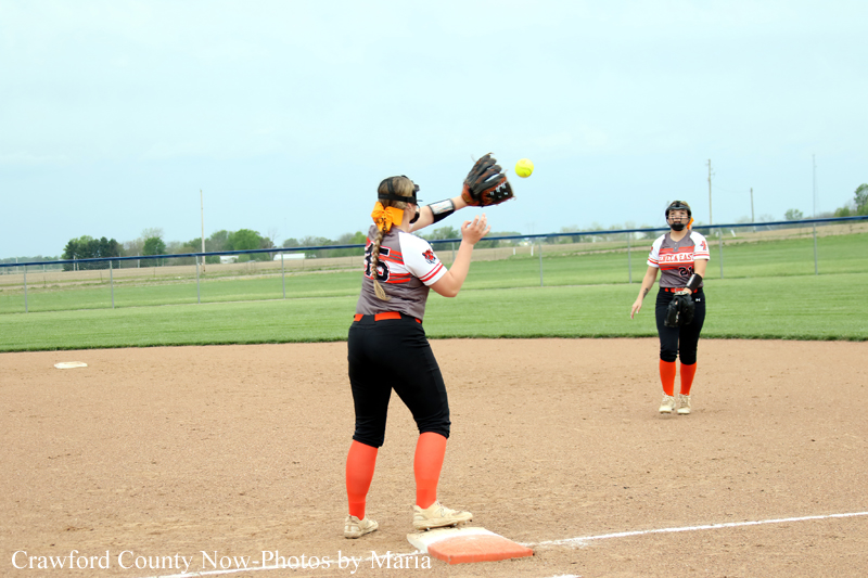 Female softball players on a infield; foreground player catches a ball near first base with glove, another player watches in the background, sunny day