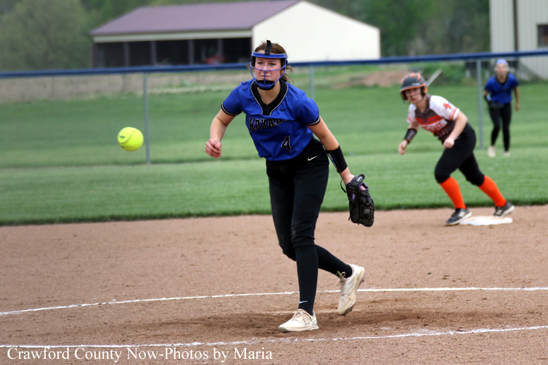 Female softball pitcher in a blue uniform on the mound, mid-pitch with a ball in the air; opponent in orange socks in the background.