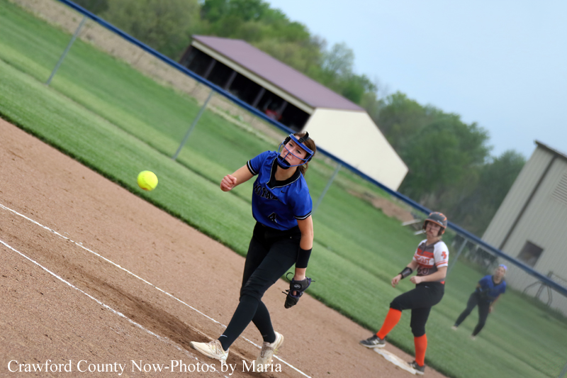 Softball pitcher in a blue uniform delivers a pitch on the dirt infield, with teammates and a field fence in the background.