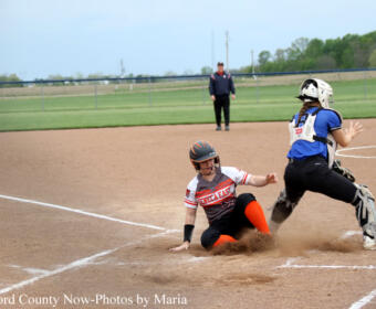 Softball action: a Seneca East runner slides into home plate as the catcher in blue gear prepares to tag her; dust rises on the infield with an umpire in the background.