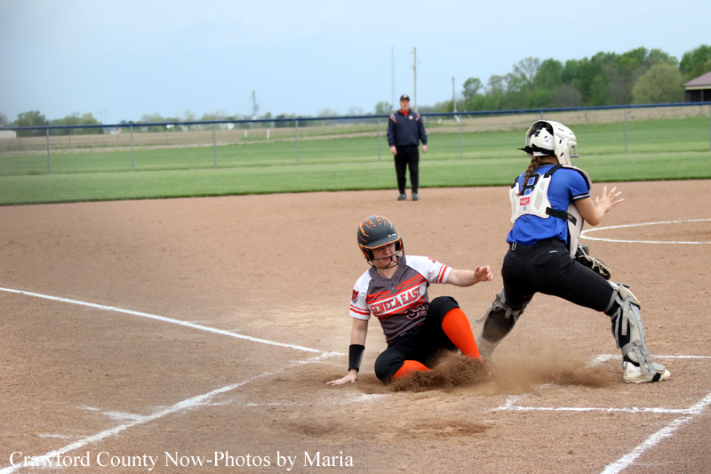 Softball action: a Seneca East runner slides into home plate as the catcher in blue gear prepares to tag her; dust rises on the infield with an umpire in the background.