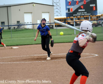 Female softball pitcher in blue winds up to throw, batter in gray and orange swings at the plate with a catcher unseen, infield dirt and scoreboard in the background.