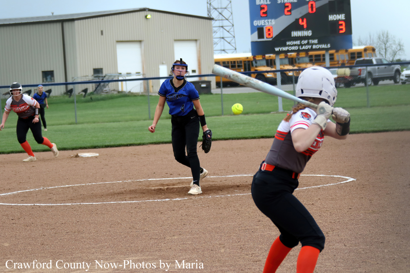 Female softball pitcher in blue winds up to throw, batter in gray and orange swings at the plate with a catcher unseen, infield dirt and scoreboard in the background.