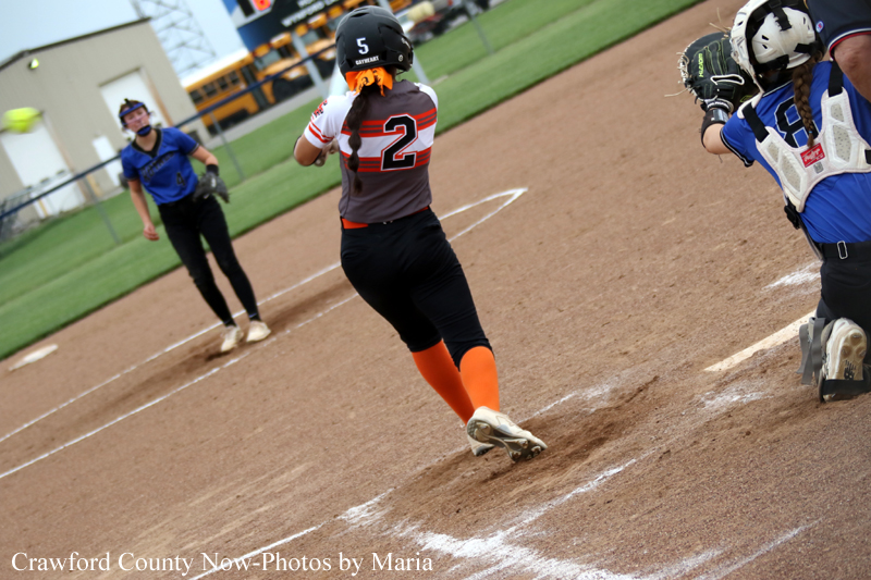 Female softball batter in gray and orange with number 2 runs toward home plate as catcher and umpire watch on a dirt field.