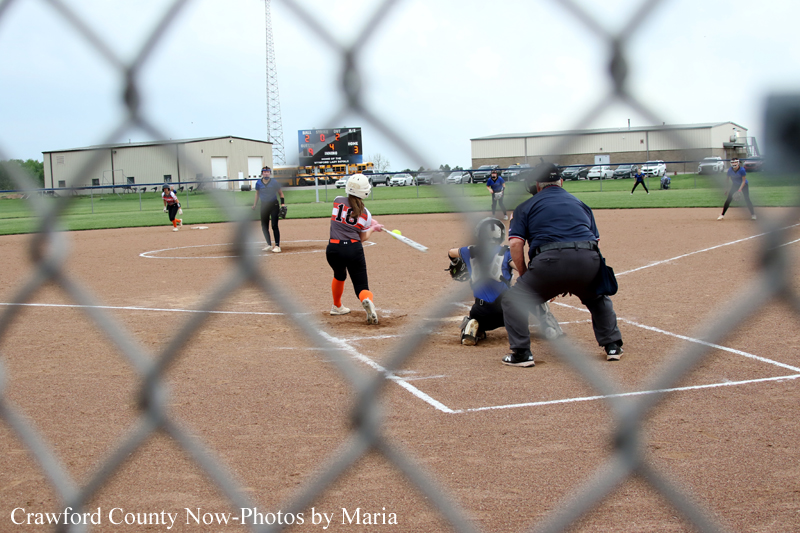 Youth softball game seen through a chain-link fence; batter swings at a pitch near home plate while catcher, umpire, and players look on in the infield with a scoreboard in the background.