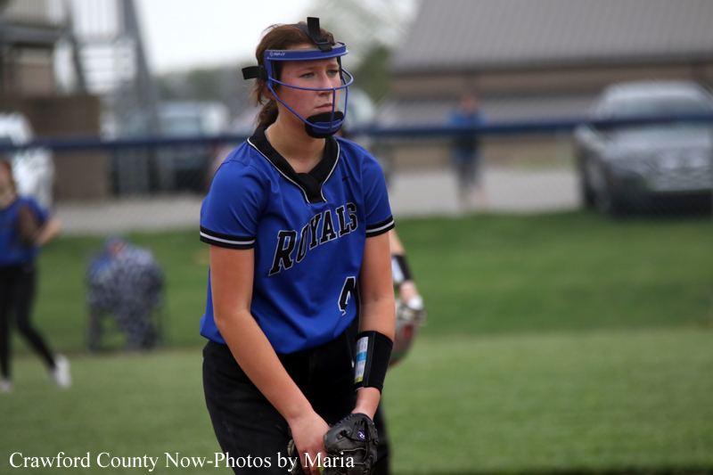 Female softball player in a blue Royals uniform stands on the field, wearing a protective helmet and glove, focused on the ball.