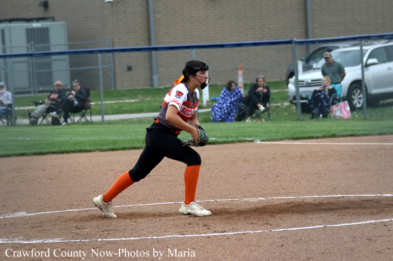 Female softball player in black pants and orange socks crouches to field a ground ball on a dirt infield, spectators behind a chain-link fence.