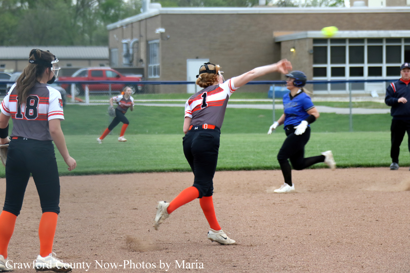 Softball game in progress: pitcher in gray-red uniform throws the ball as a blue-clad batter runs toward first base on a sunny field.