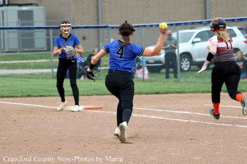 Softball pitcher in a blue jersey with number 4 throws the ball from the pitcher's mound as teammates and a runner watch on the dirt infield.