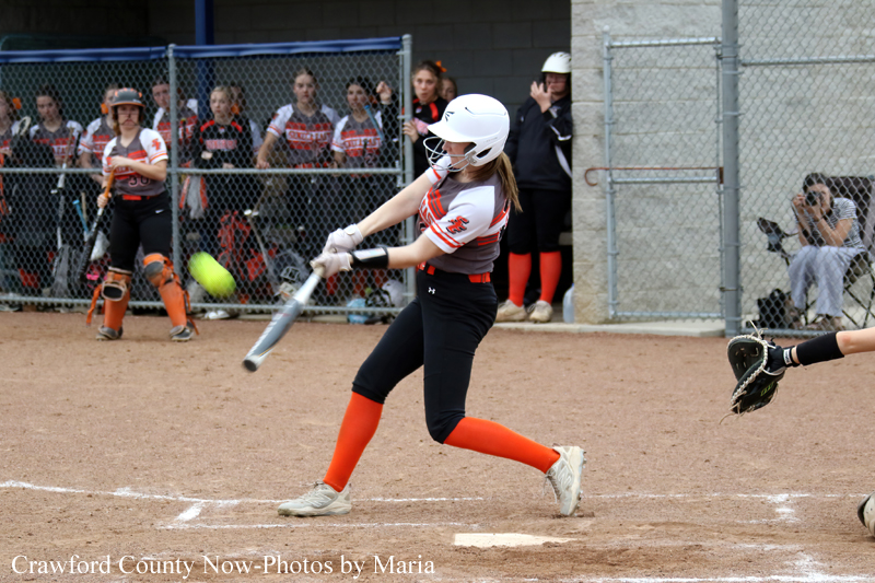 Female softball player in a white helmet swings at a pitched ball on a dirt field; spectators watch behind a chain-link fence.