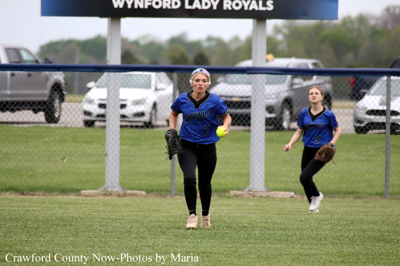 Two young softball players in blue uniforms stand on a grassy field near a chain-link fence, ready for play.