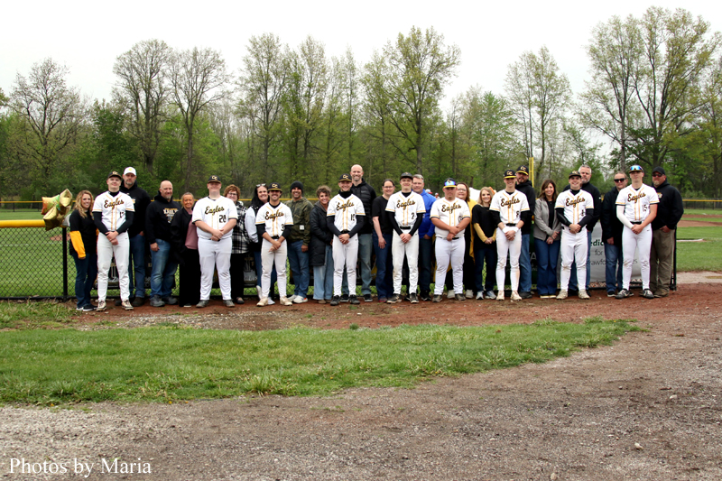 Group of baseball players in white uniforms posing with fans along a chain‑link fence on a dirt field, with trees in the background.