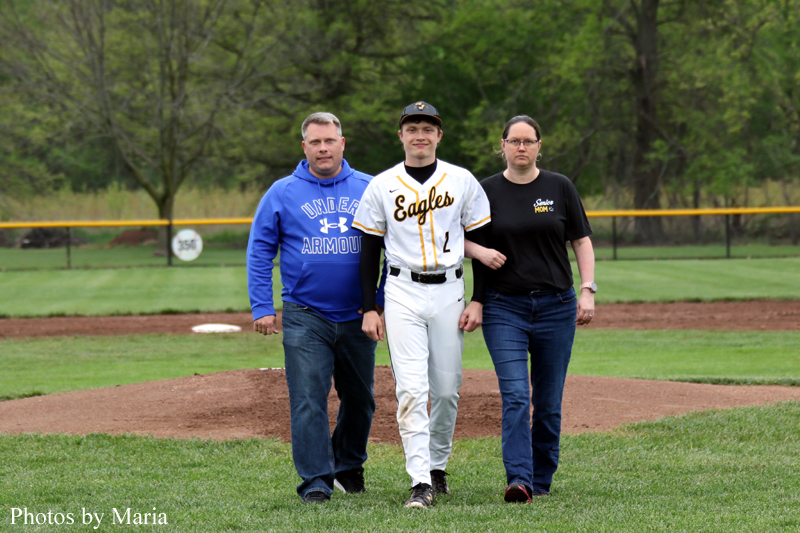Baseball player in a white Eagles uniform walks between two adults on a grassy field with a yellow outfield fence in the background.