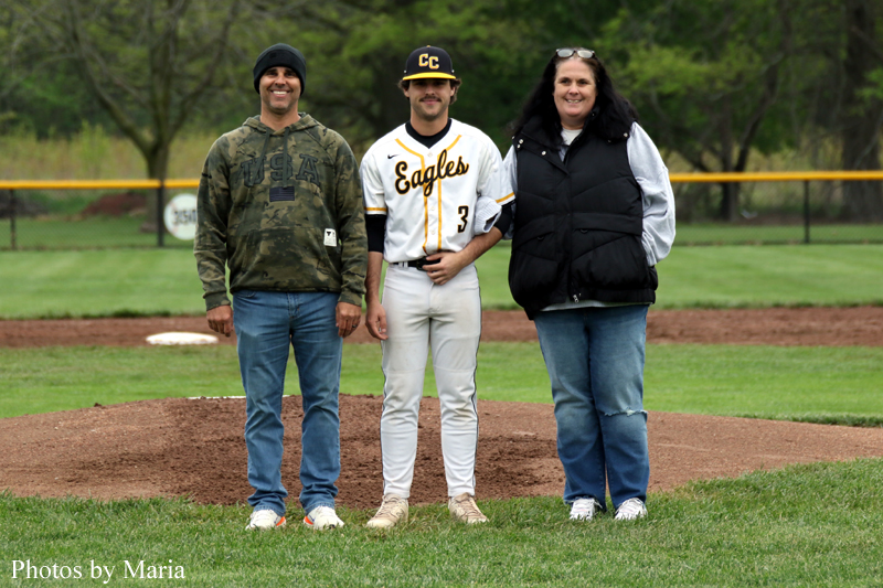 Three people pose on a baseball field: a player in a white Eagles uniform flanked by a man in a camo hoodie and a woman in a black vest.