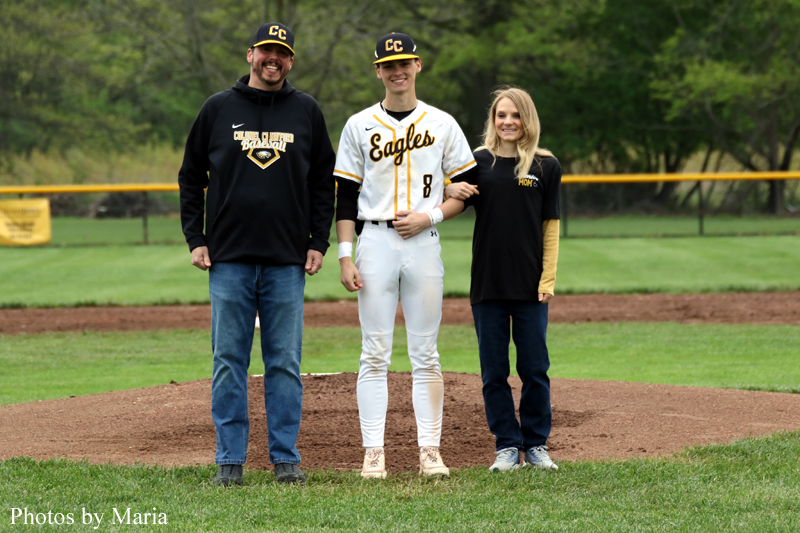 Baseball player in white and gold Eagles uniform standing between a man in a black hoodie and a woman in a black and gold shirt on the pitcher's mound.