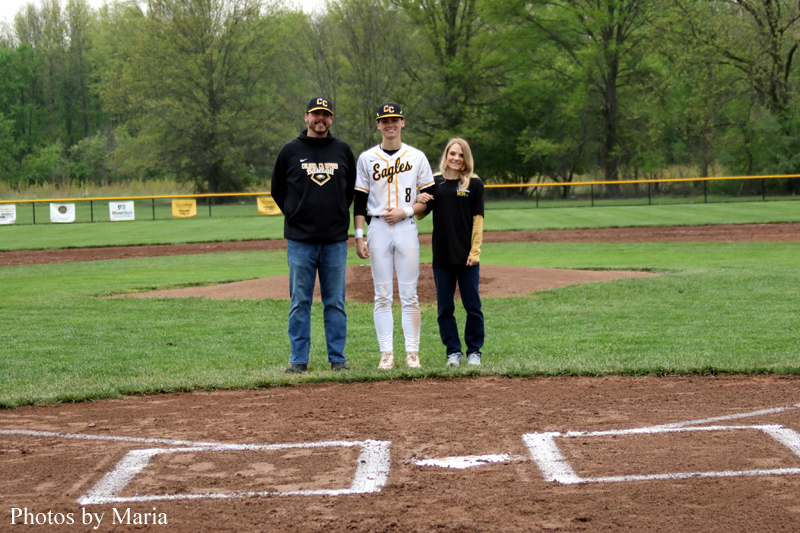 Three people stand on a baseball infield near home plate: a player in a white Eagles uniform flanked by two adults in black shirts.