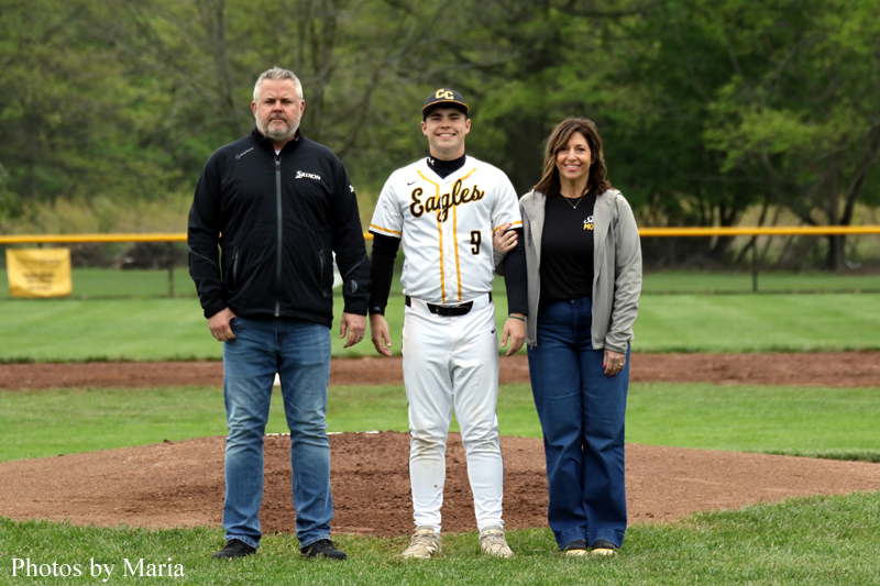 Baseball player in white and yellow Eagles uniform (#9) stands between a man in a black jacket and a woman in a gray cardigan on the pitcher's mound.