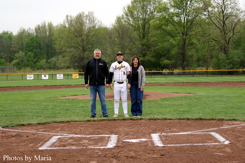 Three people pose on a baseball field near home plate; a player in a white Eagles uniform stands between two adults with trees in the background.