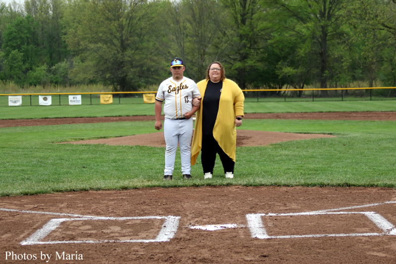 Two people stand on a baseball infield near home plate: a batter in Eagles uniform and a woman in a yellow cardigan posing together.