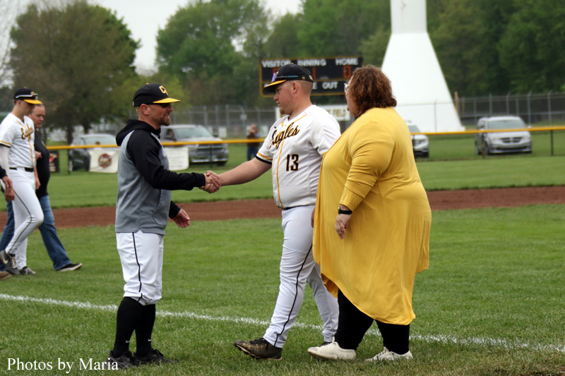 Baseball player in a white uniform (number 13) shakes hands with a man in a gray jacket on the field, while a woman in a yellow cardigan stands nearby.
