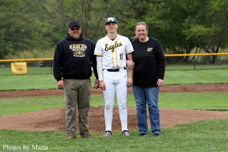 Young baseball player in a white Eagles uniform stands on a pitcher's mound, flanked by a man and a woman on a grassy field.