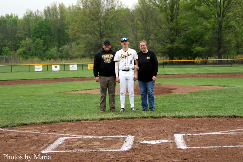 Three people stand together on a baseball field: a player in white uniform flanked by two men in dark tops, posing for a photo with the outfield and trees in the background.