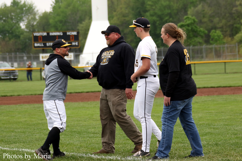 Baseball players shake hands on a diamond after a game, with an umpire/coach in a black hoodie nearby and a scoreboard in the background.