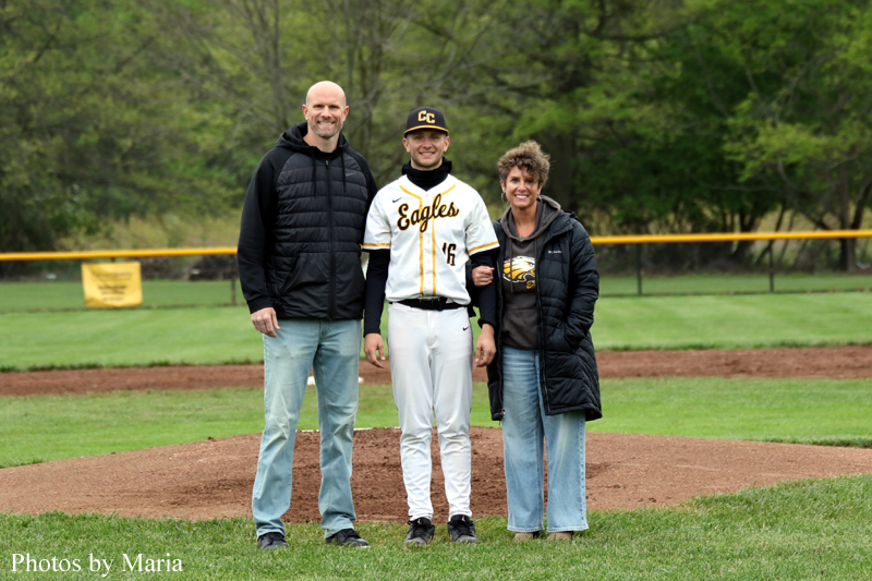 Baseball player in a white Eagles uniform standing between two adults on a grass field with a dirt mound behind them, smiling.