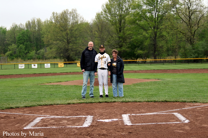 Three people pose together on a baseball infield near home plate, with a player in uniform flanked by two adults under a cloudy sky.