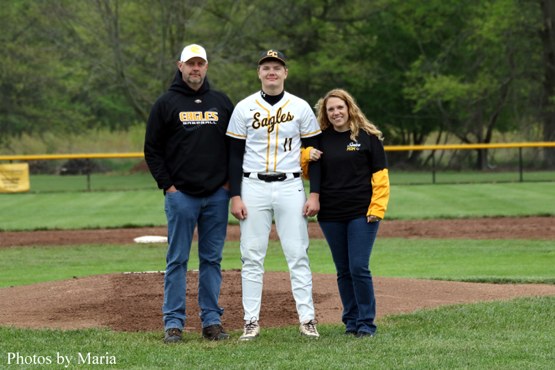Baseball player in a white and yellow Eagles uniform stands on the pitcher's mound between a man in a black Eagles hoodie and a woman in a black and yellow jacket on a baseball field.