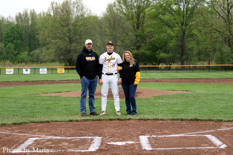 Baseball player in white and yellow uniform stands between two adults on a grassy baseball field, with the infield dirt and trees in the background.