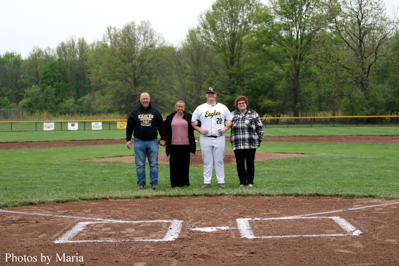 Five people stand arm-in-arm on a baseball field near home plate, with a grassy outfield and trees in the background, posing for a photo.