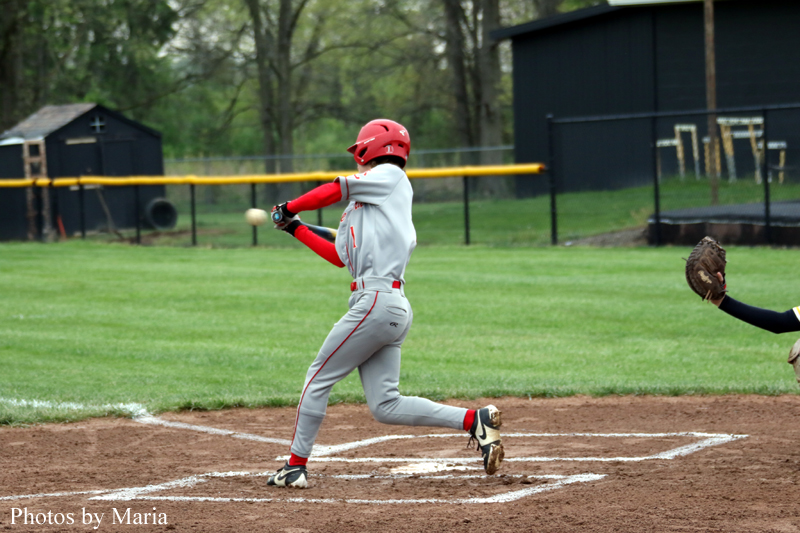Baseball batter in gray and red uniform swings at a pitch at home plate on a dirt infield urban field backdrop.