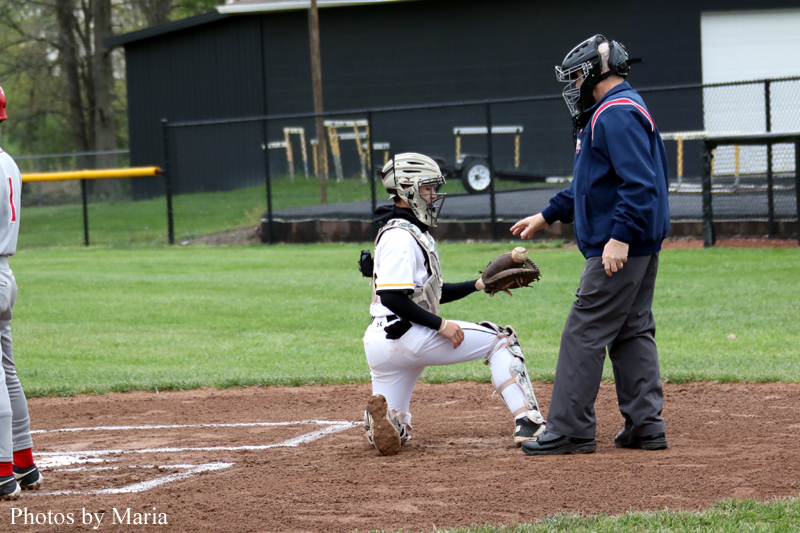 Baseball catcher kneels at home plate and talks to the umpire during a game on a dirt infield at a park field.