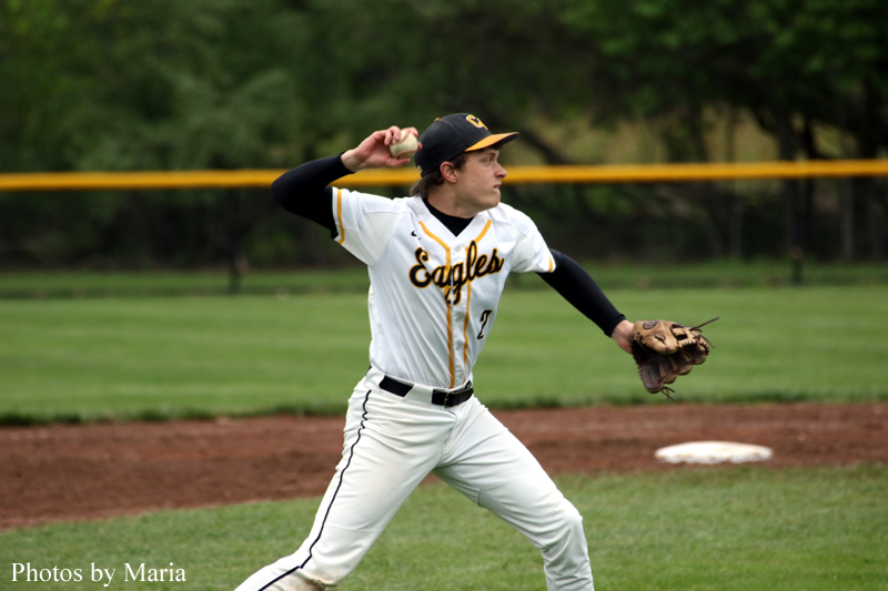 Baseball pitcher in a white Eagles uniform winds up to throw on a grassy field with a dirt infield and yellow outfield fence in the background, glove ready on his left hand.